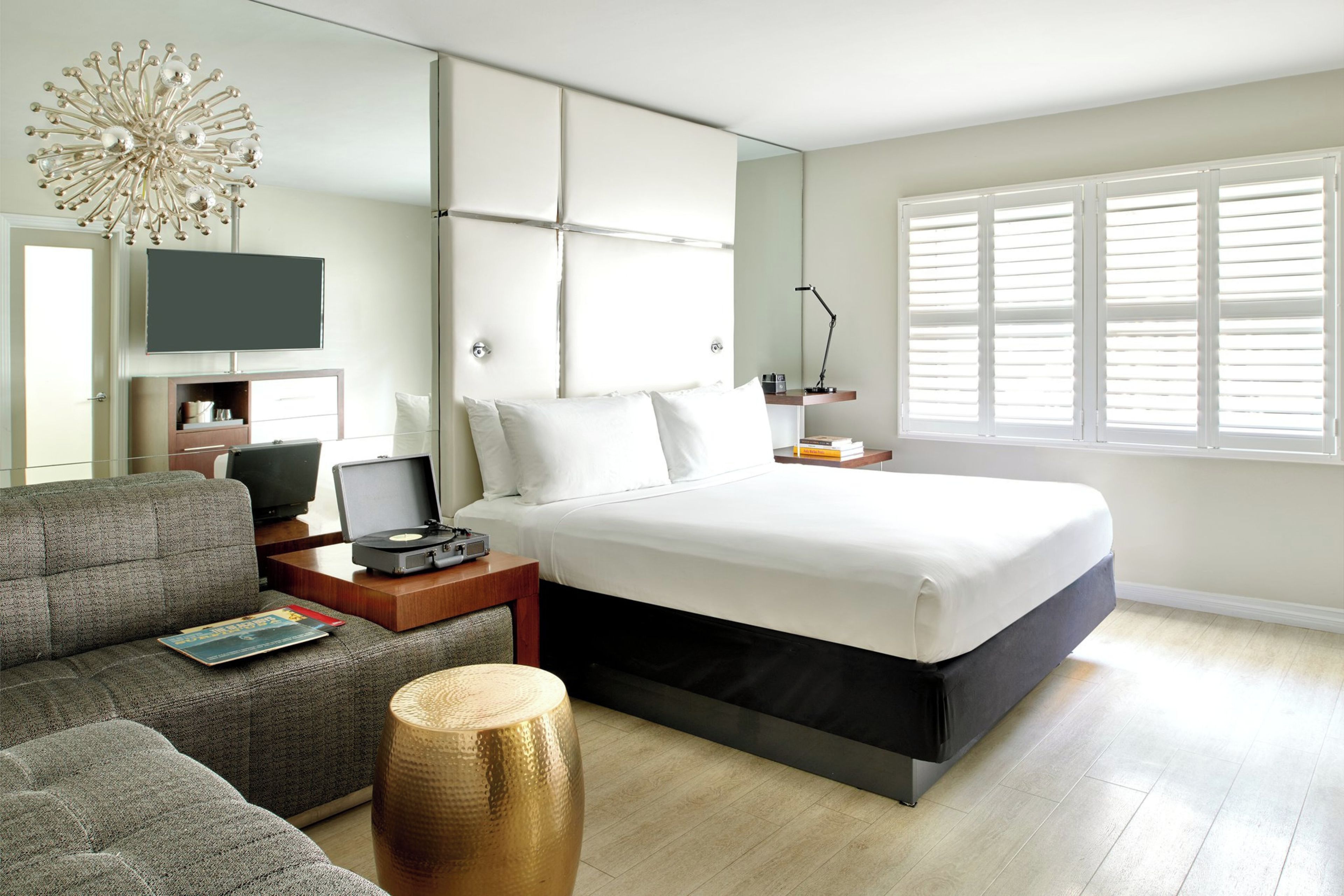 Een moderne hotelkamer met een bed, een slaapbank, een bureau met een laptop, een televisie en een groot raam met jaloezieën.