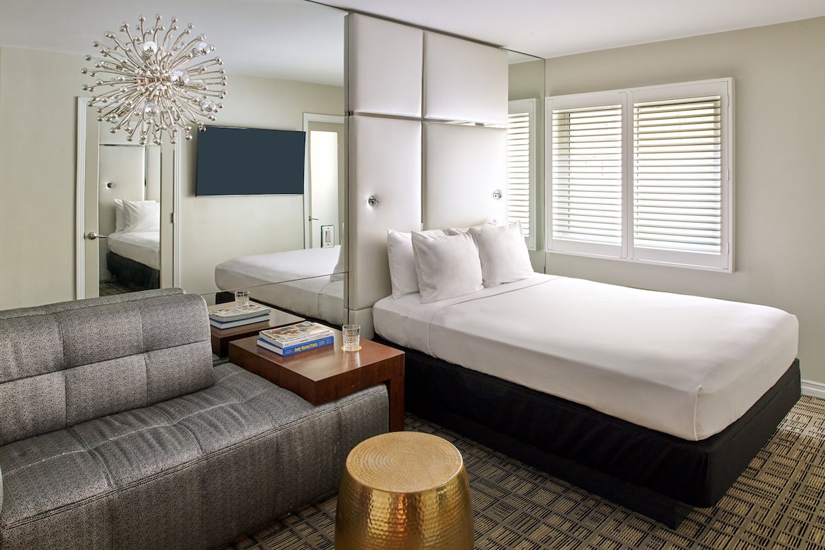 A modern hotel room with a large bed, a sofa, a bedside table with books, a decorative lamp, and a large mirror.