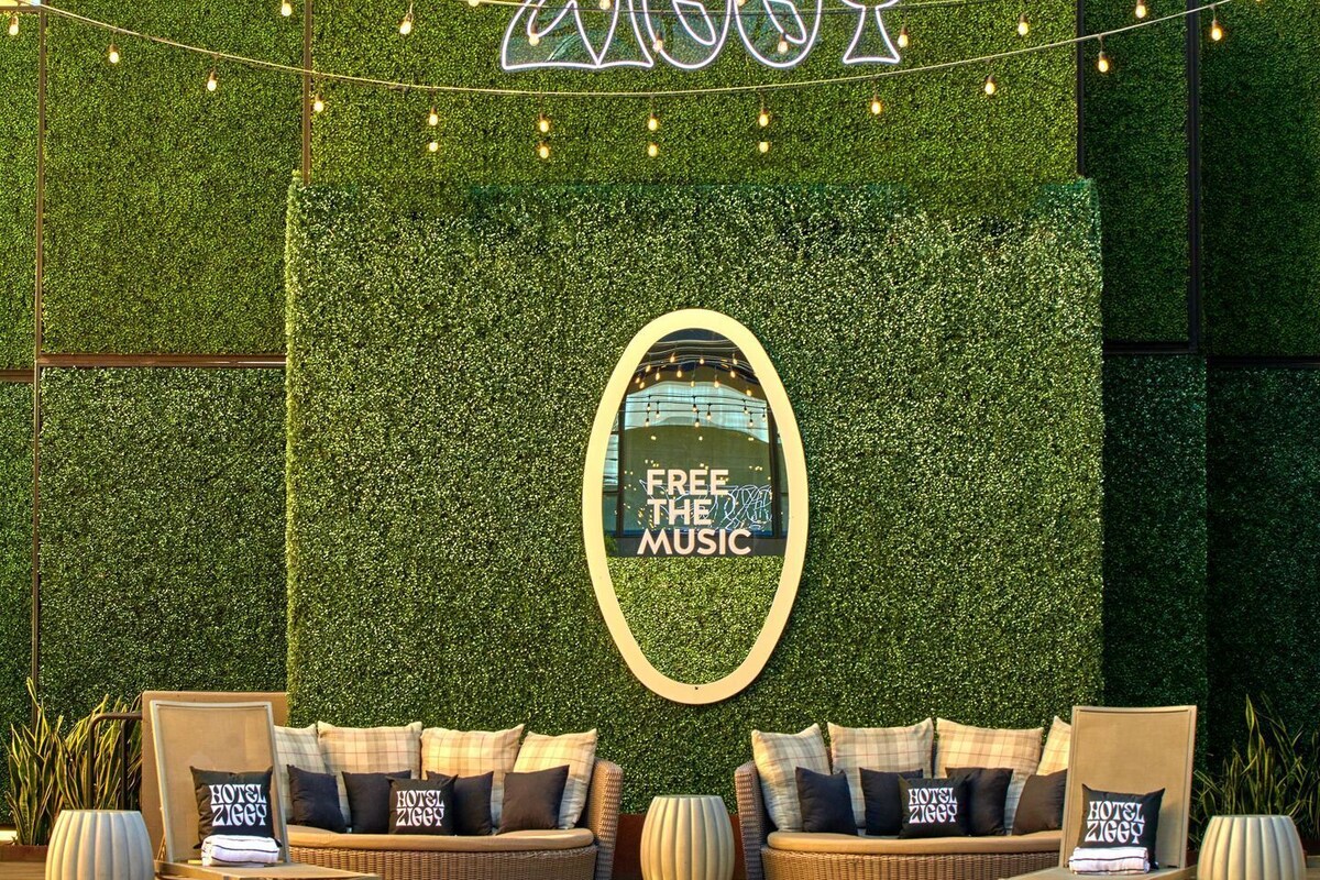 A poolside area with a green wall, a sign reading 'FREE HEAR MUSIC,' and lounge seating with pillows labeled 'HOTEL ZIGZAG.'