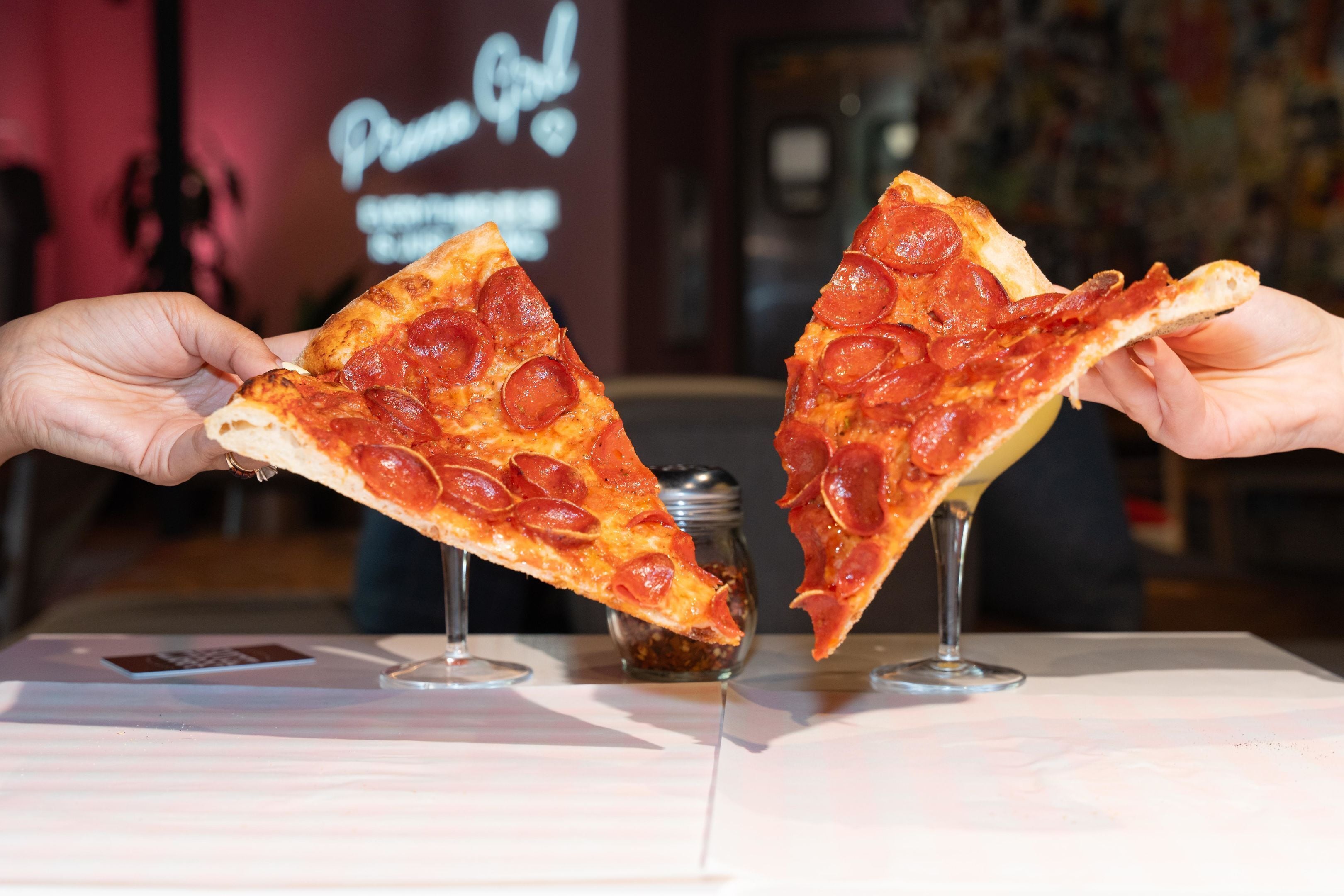 Dos manos sosteniendo rebanadas de pizza de pepperoni sobre una mesa.