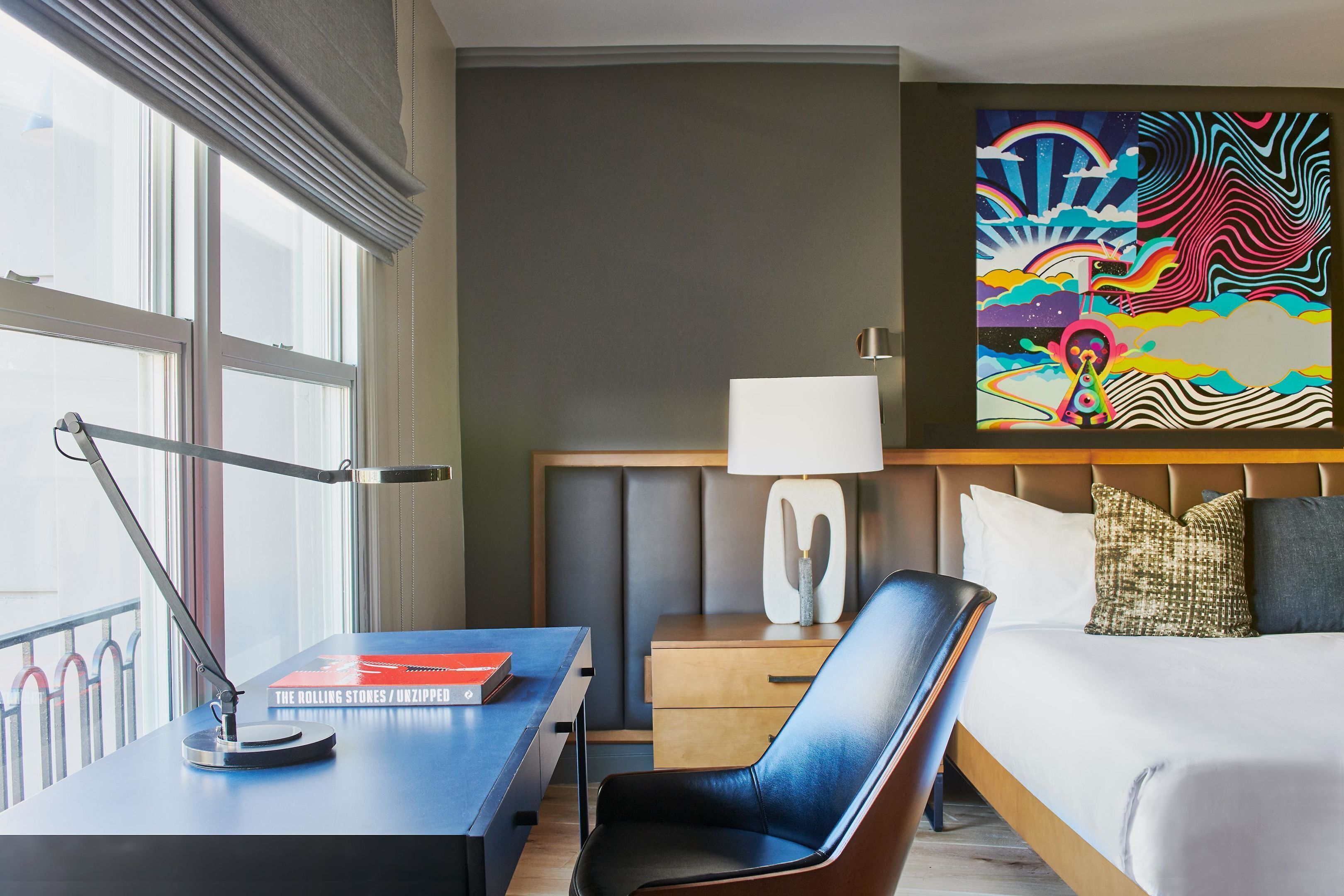 A hotel room with a desk, chair, bed, bedside table, lamp, and a large abstract painting on the wall.