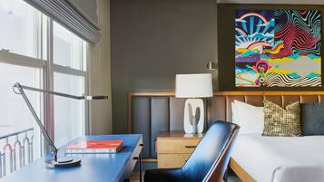 A hotel room with a desk, chair, bed, bedside table, lamp, and a large abstract painting on the wall.