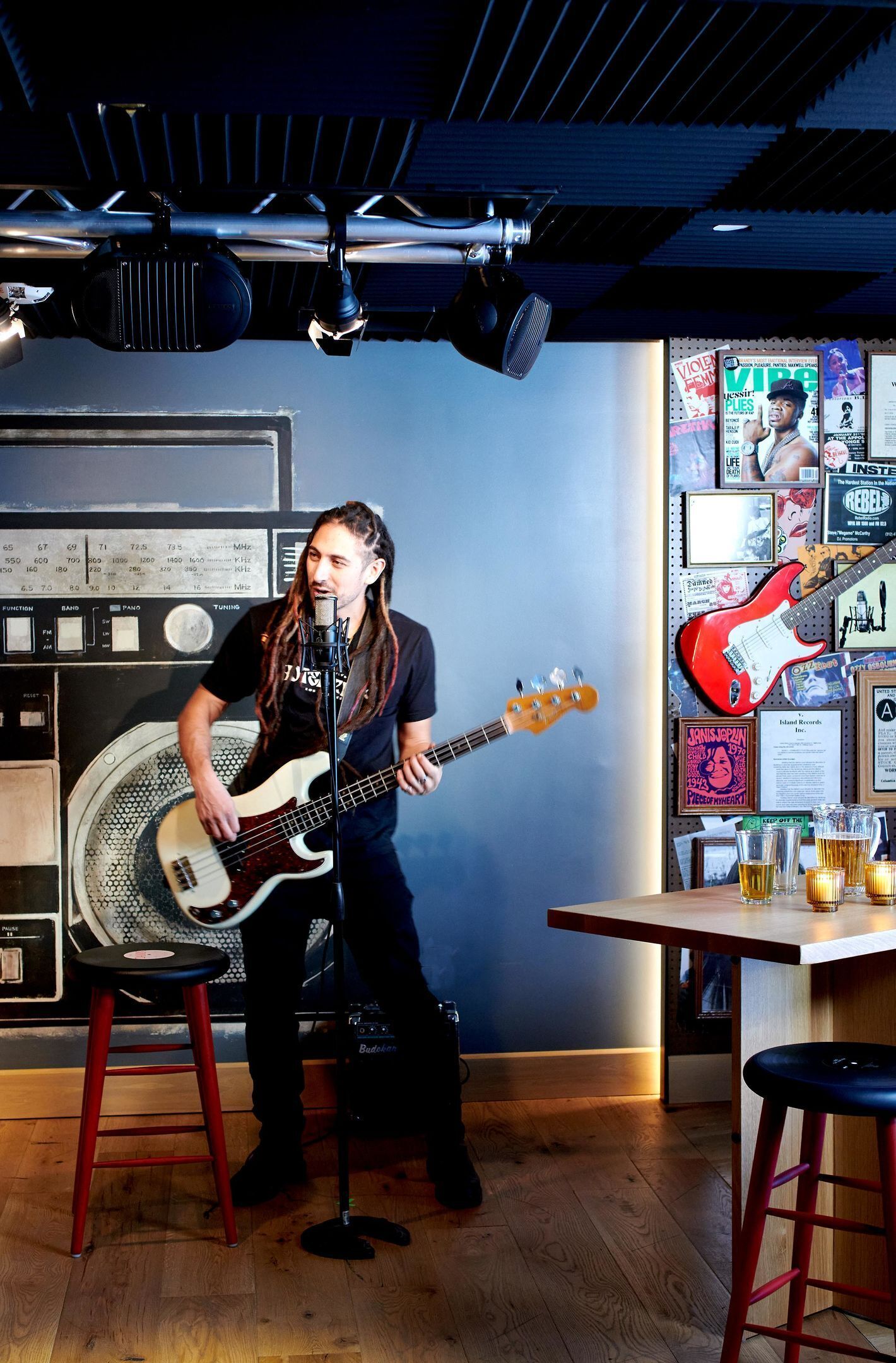Un músico tocando el bajo en una sala con una mesa de mezclas, carteles en la pared y taburetes.