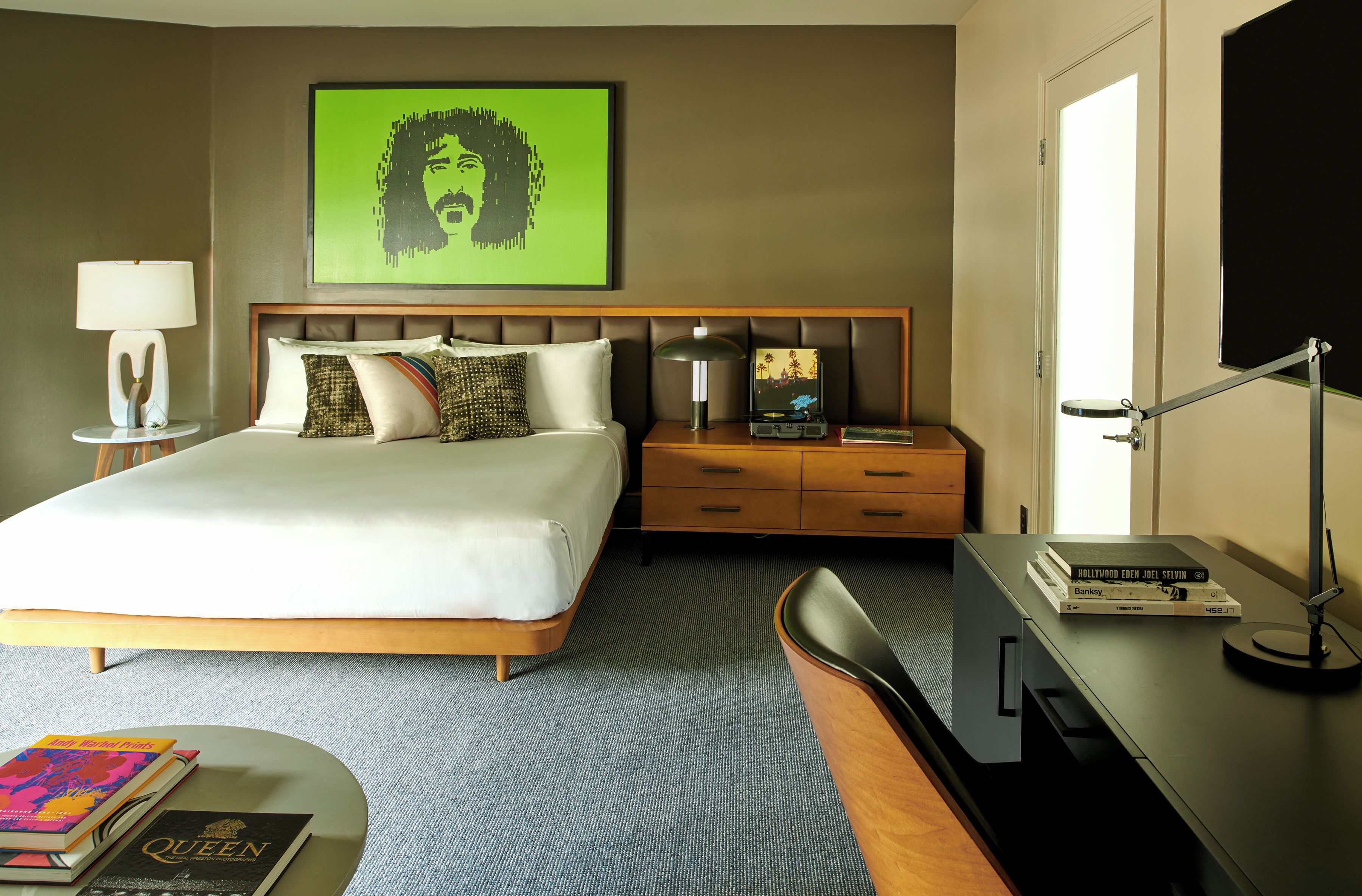 A hotel room with a bed, bedside table, lamp, and a desk with a computer and books.
