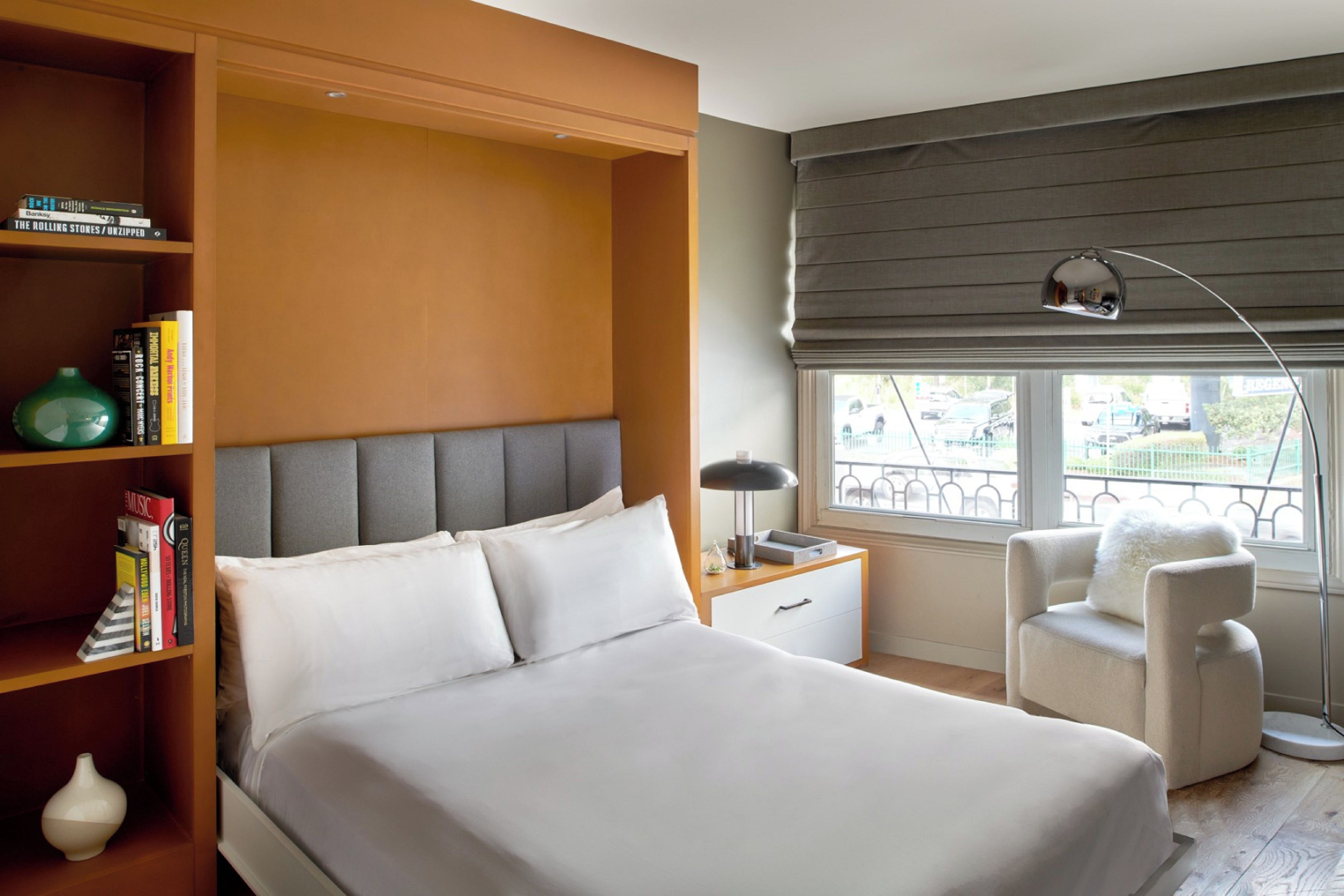 A modern bedroom with a large bed, a bedside table, a chair, a bookshelf, and a window with a view of the outside.