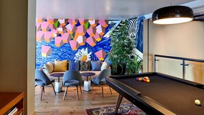 Una mesa de billar con bolas, un sofá con cojines, una mesita redonda y un mural de pared con motivos geométricos y coloridos.