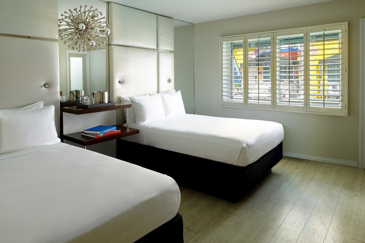A hotel room with two beds, a bedside table, a mirror, and a window with blinds.