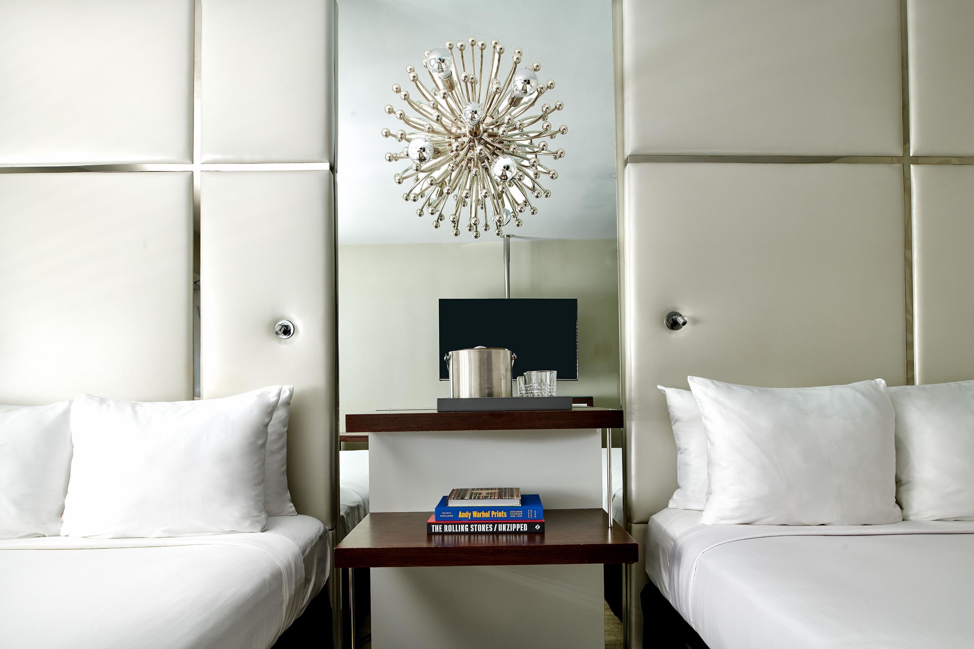 Une chambre d’hôtel avec deux lits, une table de chevet avec un livre, un luminaire décoratif et une petite étagère avec une bouilloire et des verres.