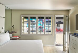 A hotel room with a bed, bedside table, lamp, and a window with blinds.