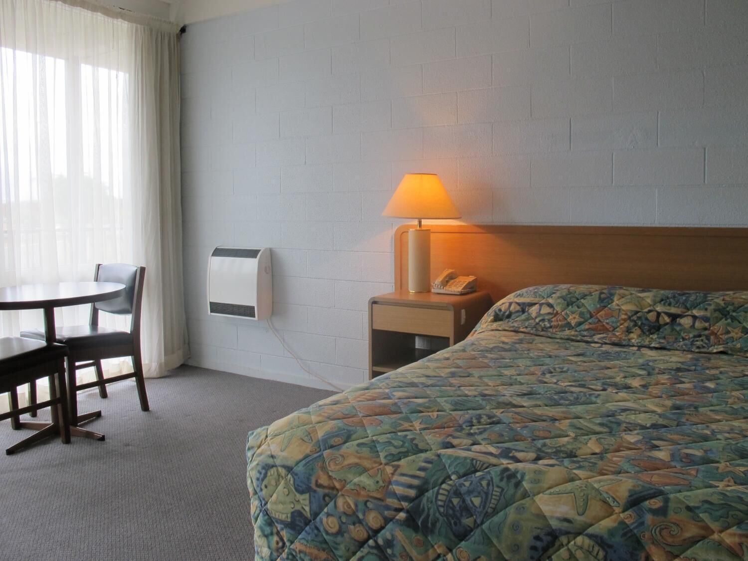 A hotel room with a bed, a bedside table with a lamp, a phone, and a wall-mounted air conditioner.