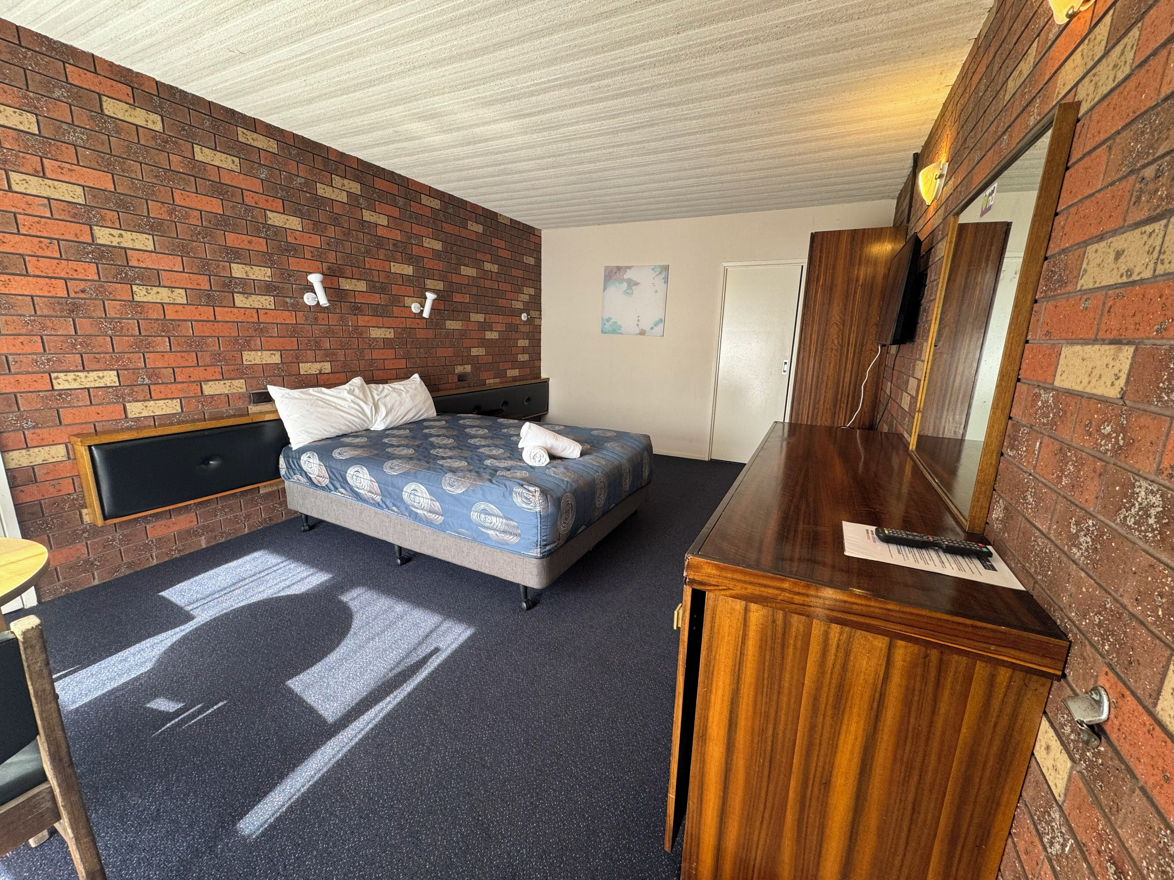 A hotel room with a brick wall, a bed with a blue patterned bedspread, a wooden dresser, and a small table.