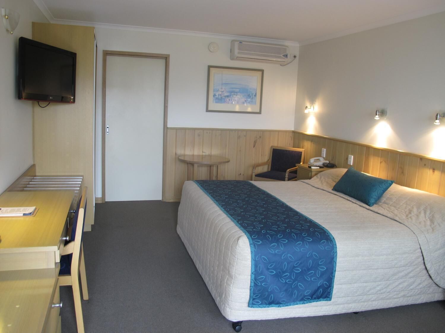 A hotel room with a bed, a desk, a chair, a television, and a painting on the wall.