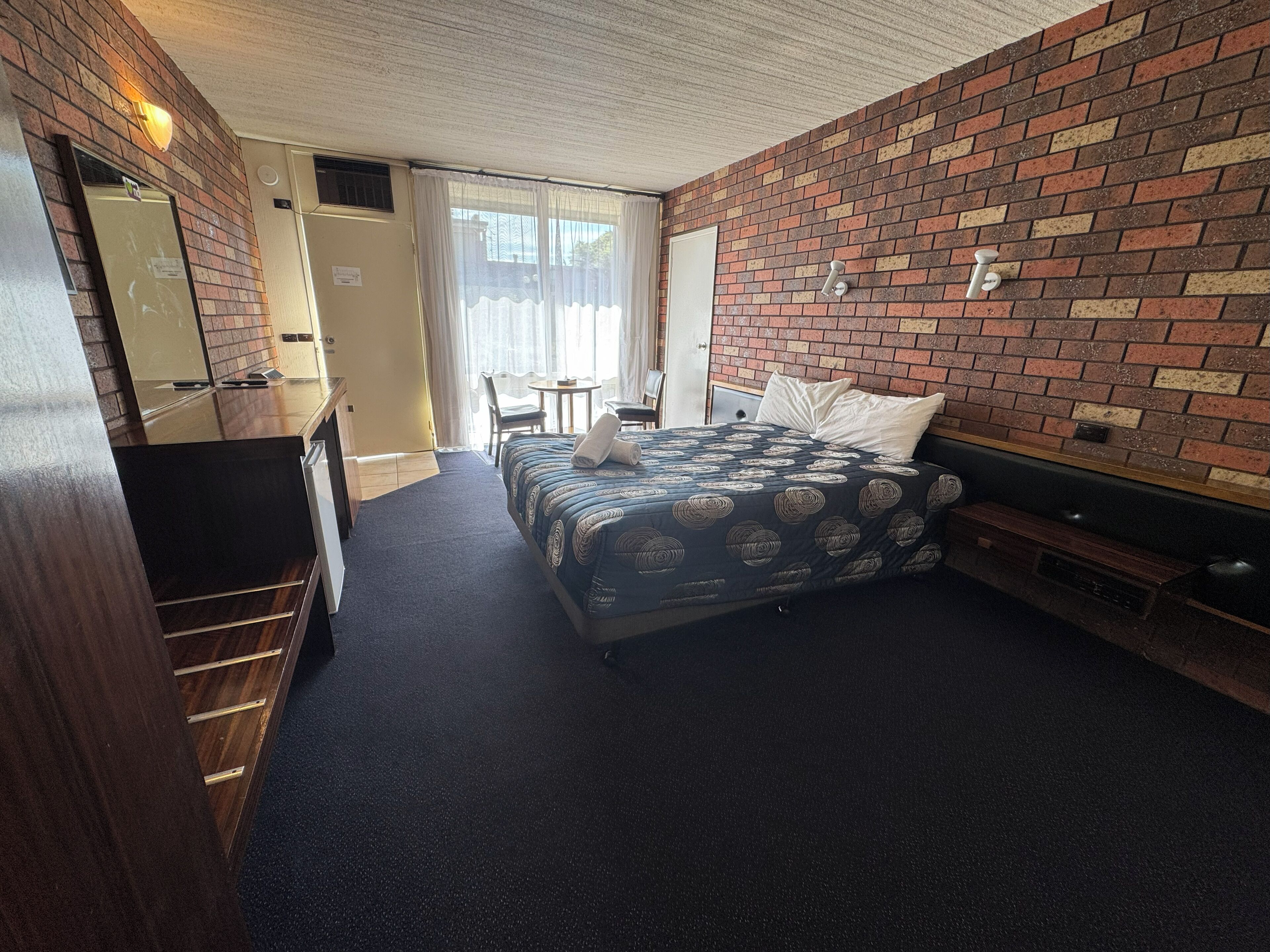 A hotel room with a bed, a desk, a chair, a small table, and a brick-patterned wall.