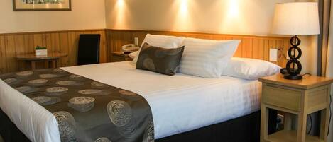 A hotel room with a large bed, a bedside table with a lamp, a small table with a plant, and a wall-mounted telephone.