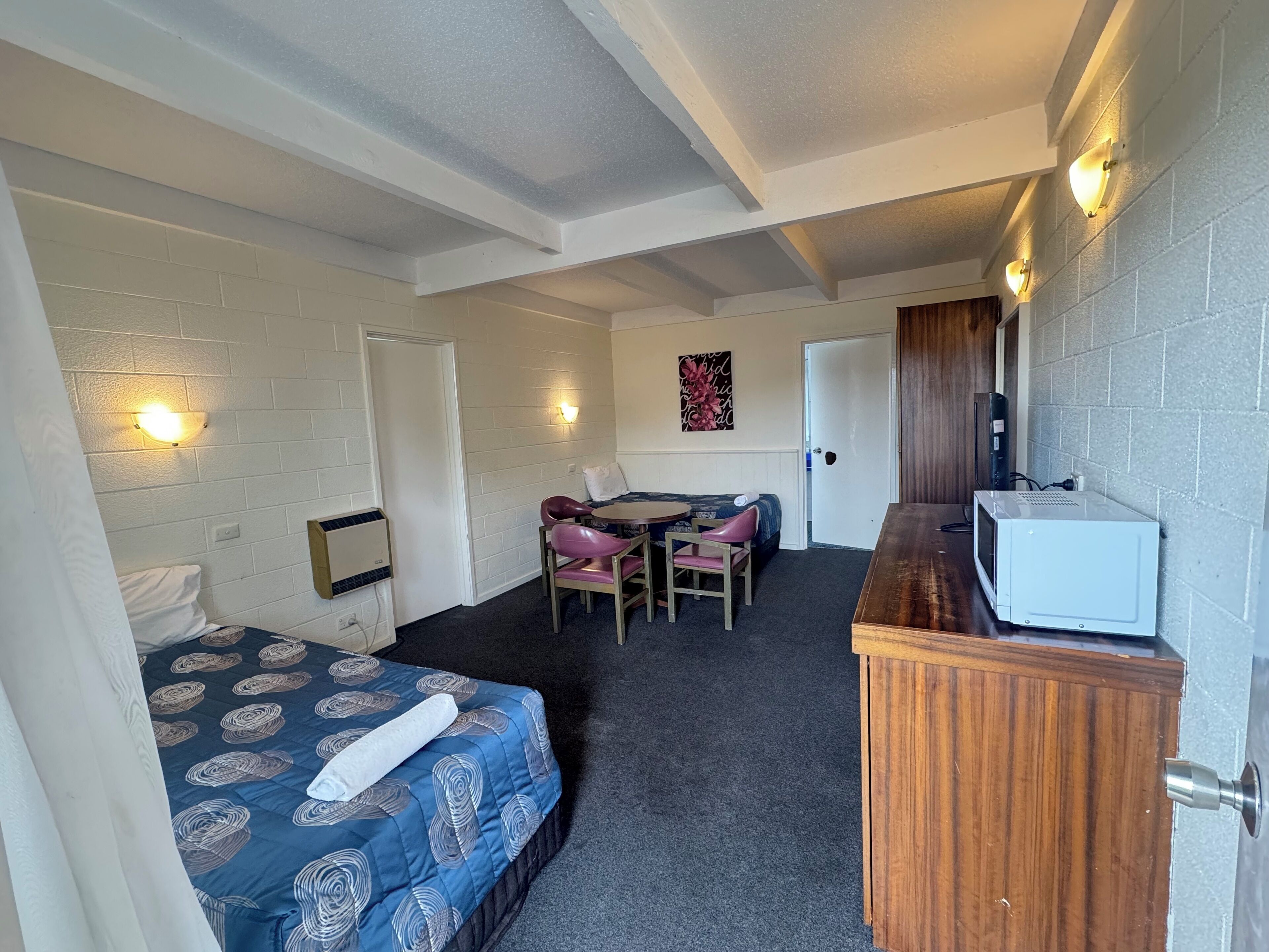 A compact hotel room with a bed, a desk with a chair, a microwave, and a wall-mounted TV.