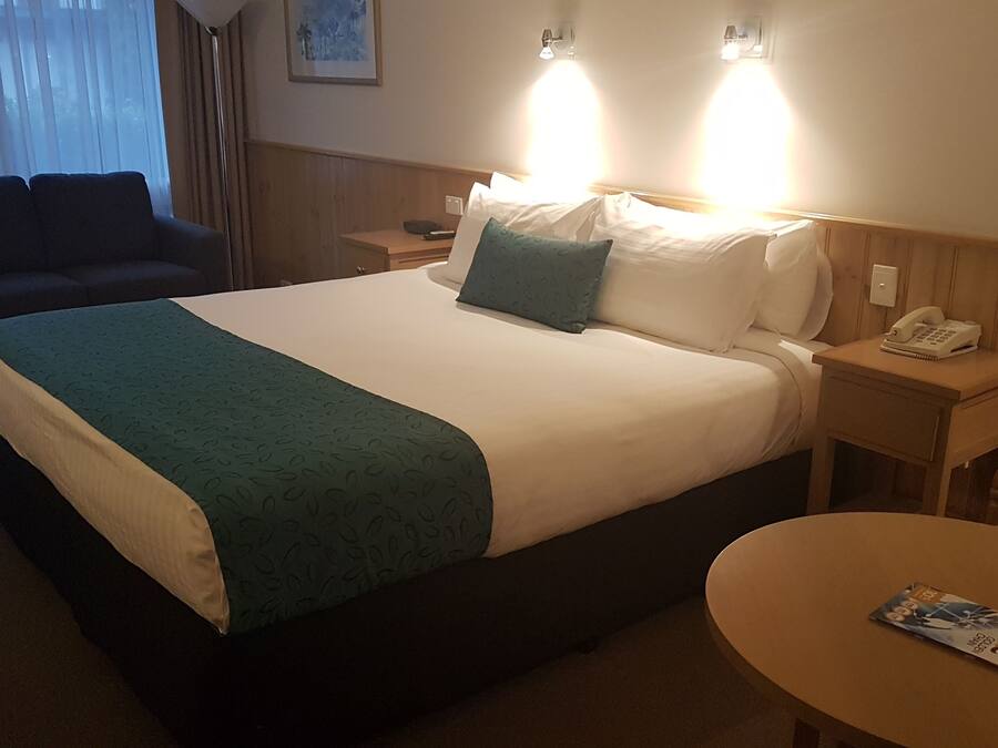 Habitación de hotel con una cama grande, una mesita de noche con un teléfono, una silla y una pequeña mesa con una revista.
