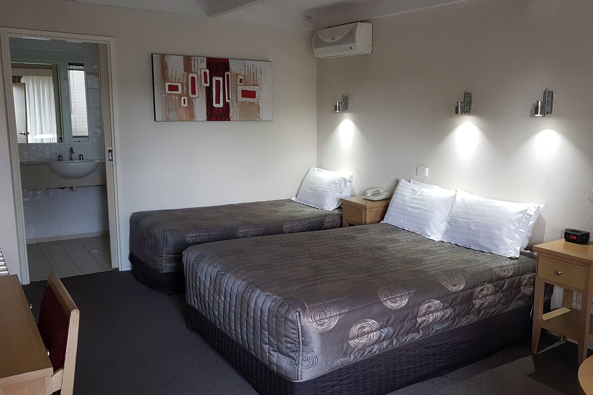 Habitación de hotel con dos camas, una mesita de noche con lámpara, un escritorio y un baño visible a través de una puerta abierta.