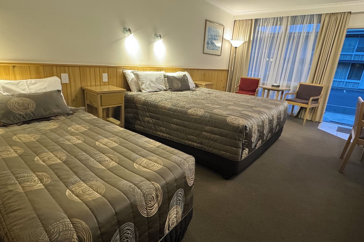 Habitación de hotel con dos camas, una mesita, una silla y vistas al exterior a través de una ventana con cortinas.