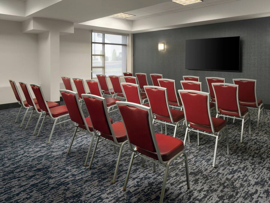 Une salle de conférence avec des rangées de chaises rouges, un grand téléviseur à écran plat accroché au mur et une moquette à motifs.