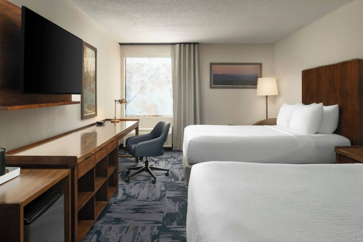 A hotel room with two beds, a desk, a chair, a TV, and a window with curtains.