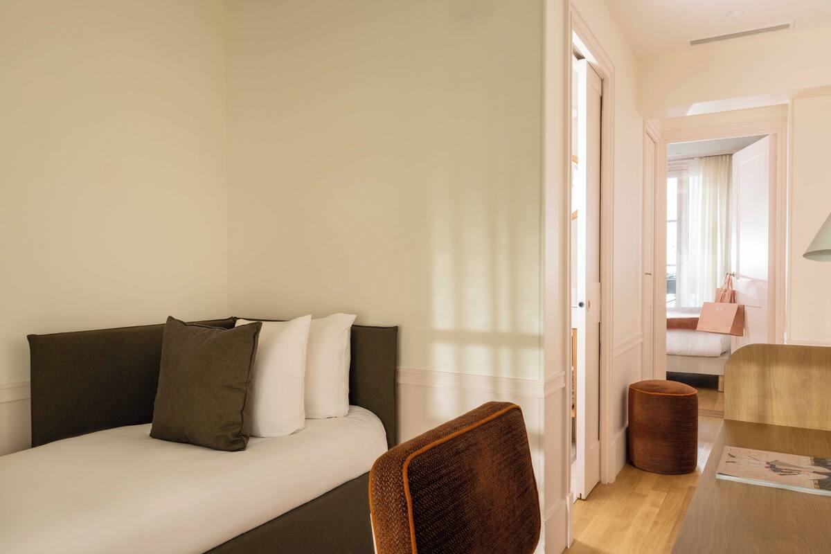 Una habitación de hotel moderna con una cama sofá, una mesita de madera y un pequeño otoman redondo.