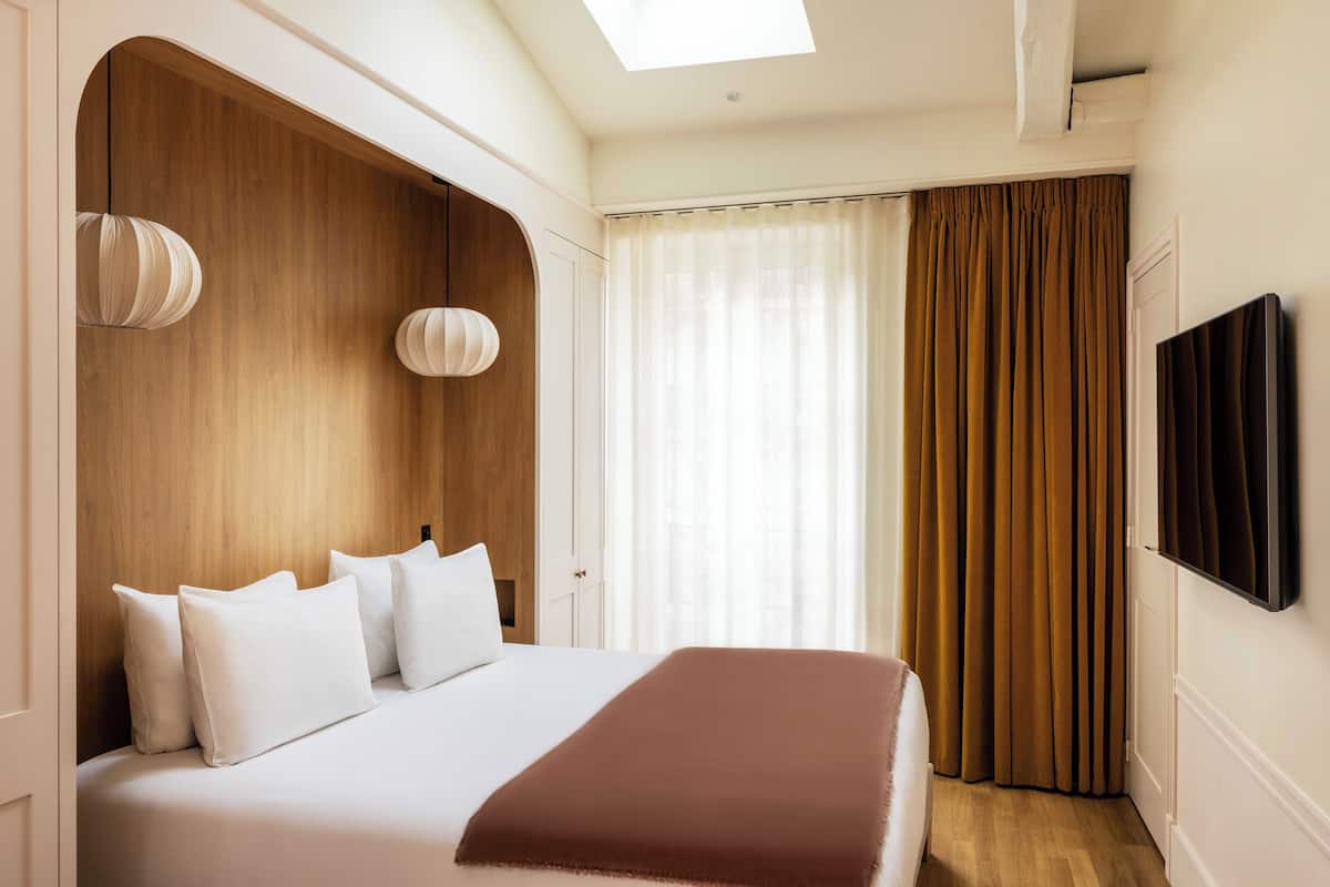 Habitación de hotel con una cama grande, cabecera de madera, televisor de pantalla plana y ventana con cortinas.