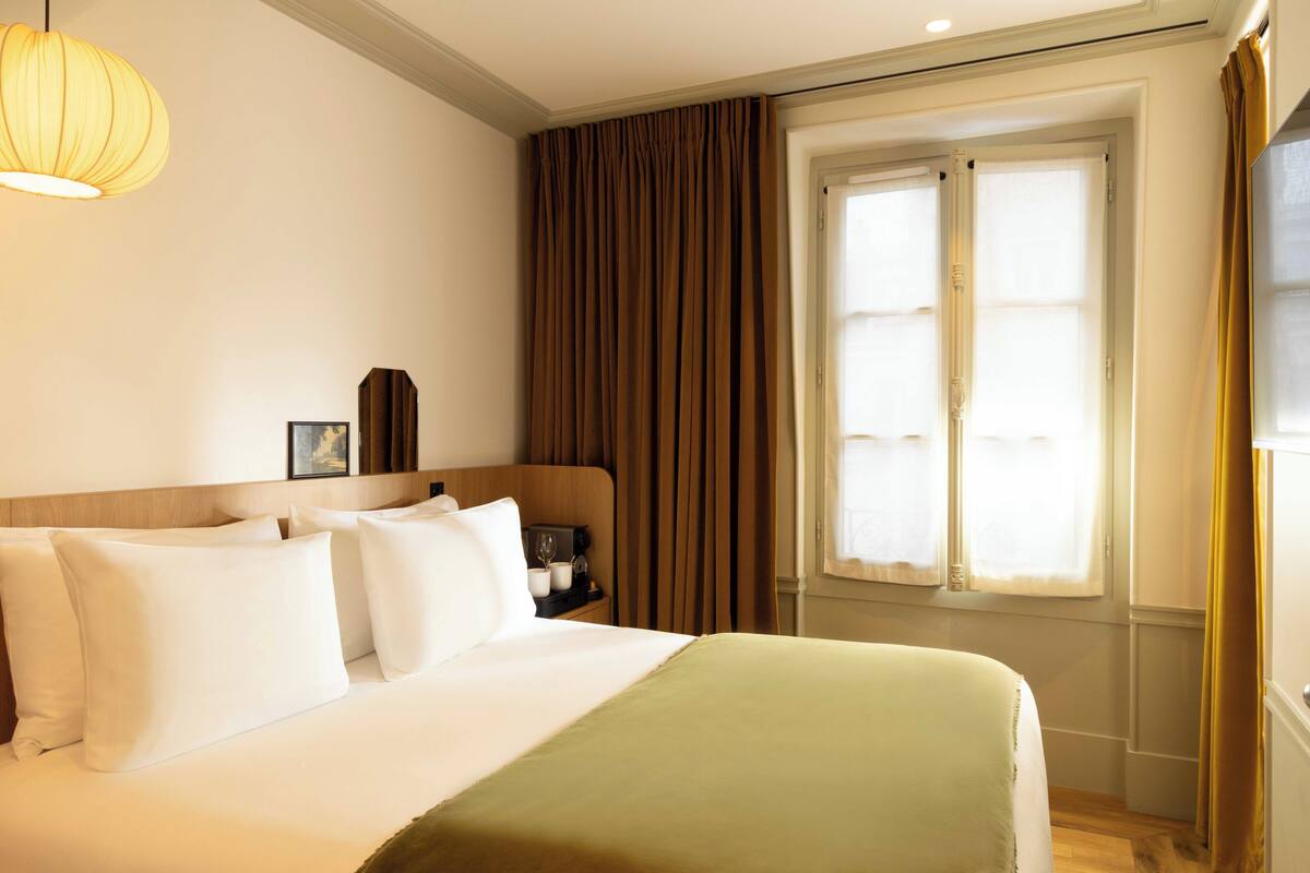 Una cama bien hecha con sábanas blancas y un plaid verde, una mesita de noche con una lámpara, y una ventana con cortinas.