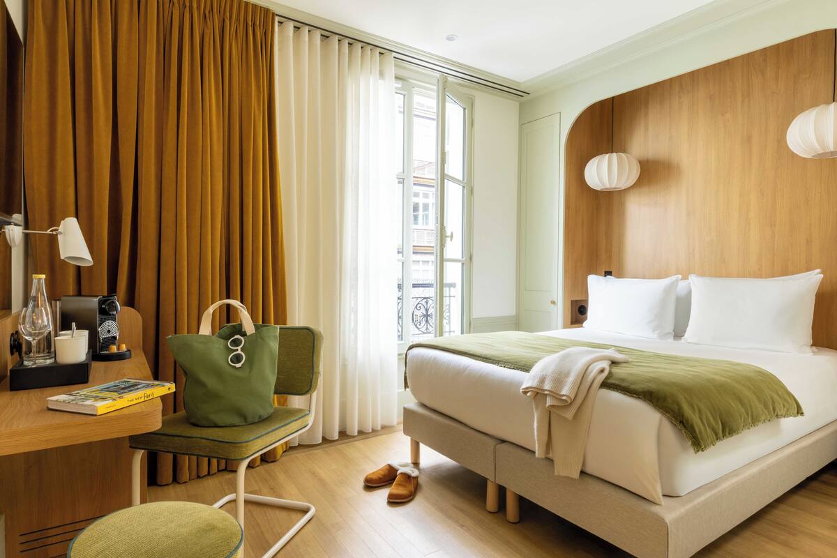 Una habitación de hotel con cama, escritorio con lámpara, silla, bolso verde y ventana con cortinas.
