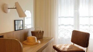A room with a wooden desk, a wicker basket, a wicker hat, a brown leather chair, a wall-mounted lamp, and a window with sheer curtains. - Hotel Casimir (Paris)