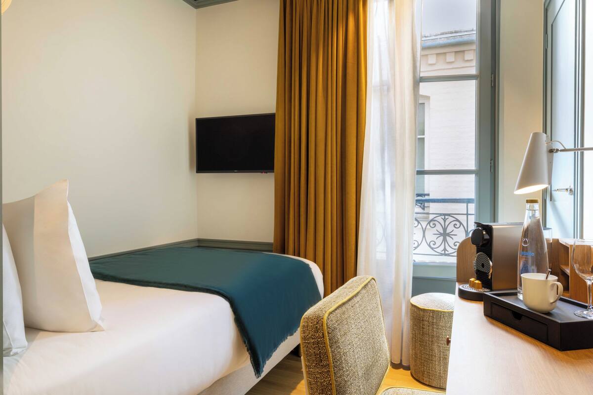 Una habitación de hotel con cama, una silla, cafetera, taza y televisor en la pared.