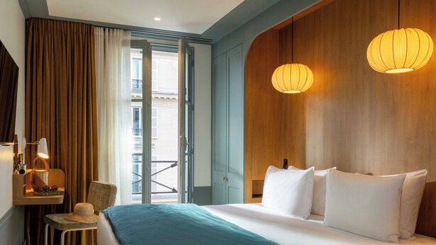 A hotel room with a bed, a chair, a desk with a lamp, and a view of the outside balcony. - Hotel Casimir (Paris)
