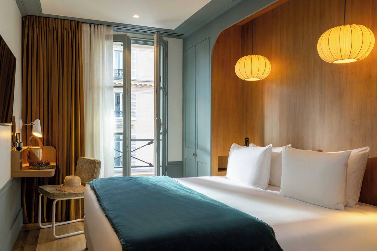 Habitación de hotel con cama, una silla, escritorio con lámpara y vista al balcón exterior.