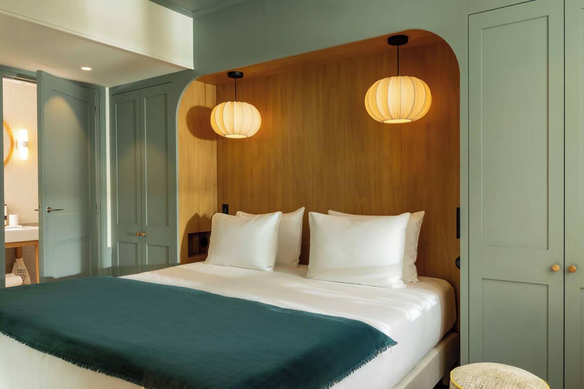 Una cama bien hecha con sábanas blancas y una manta color turquesa, cabecera de madera con dos lámparas colgantes, y una pequeña silla redonda con asiento tejido.