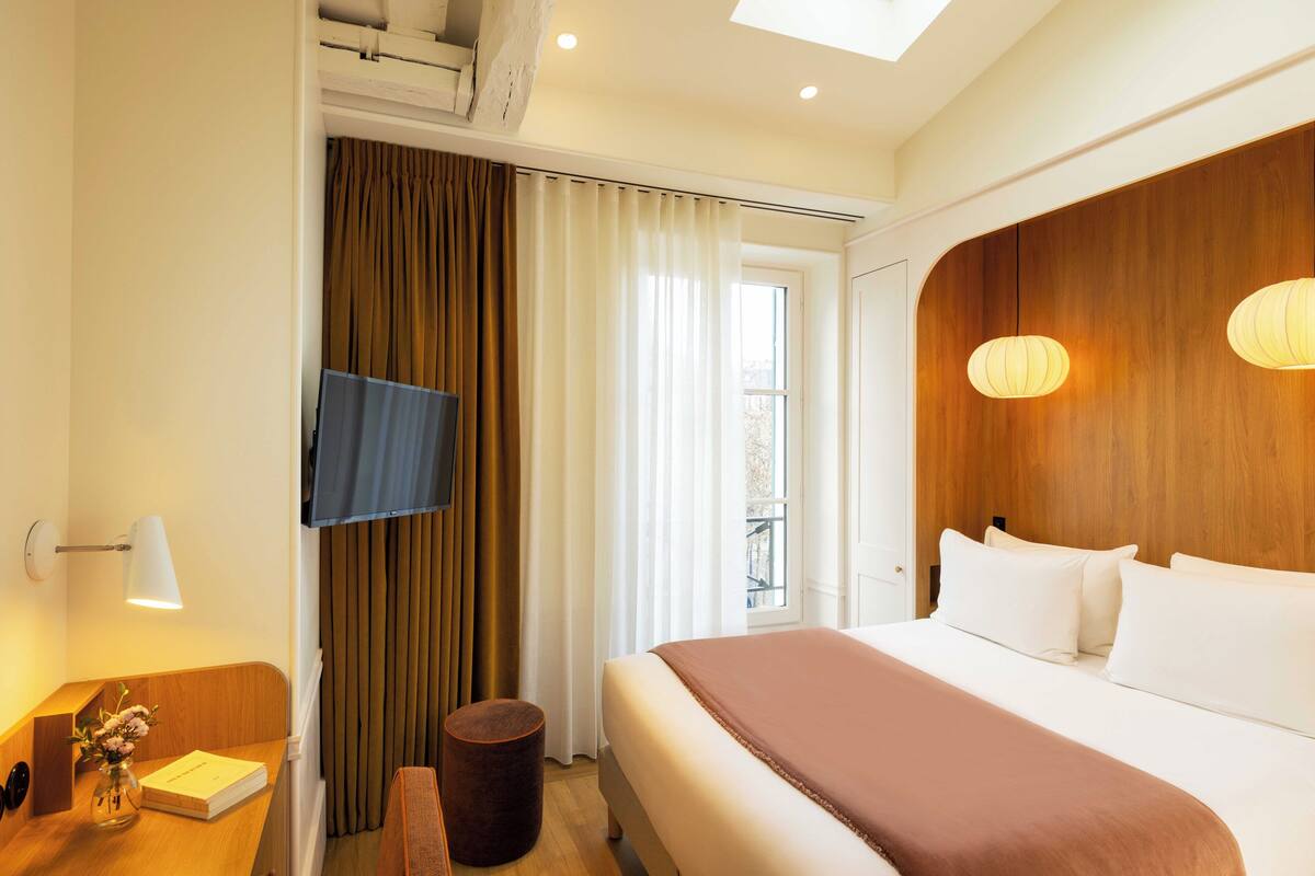 Habitación de hotel con cama, escritorio con lámpara, televisor y vistas al exterior.