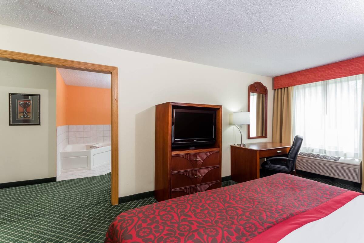 Une chambre d’hôtel comprenant un lit, une télévision, un bureau, une chaise, une salle de bain avec une baignoire et une grande fenêtre avec des rideaux.