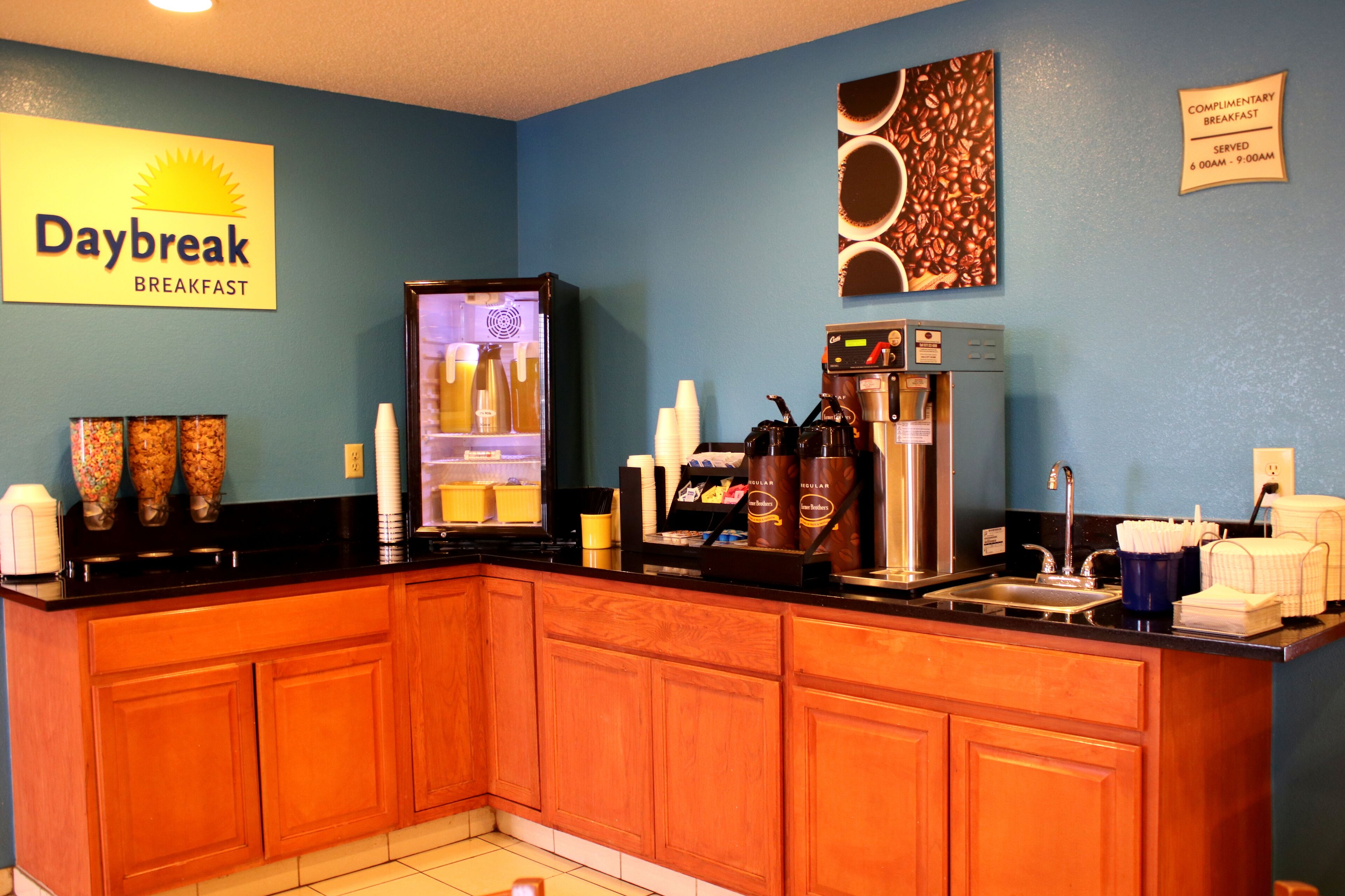 A breakfast area with a coffee machine, cups, and a sign indicating complimentary breakfast hours.