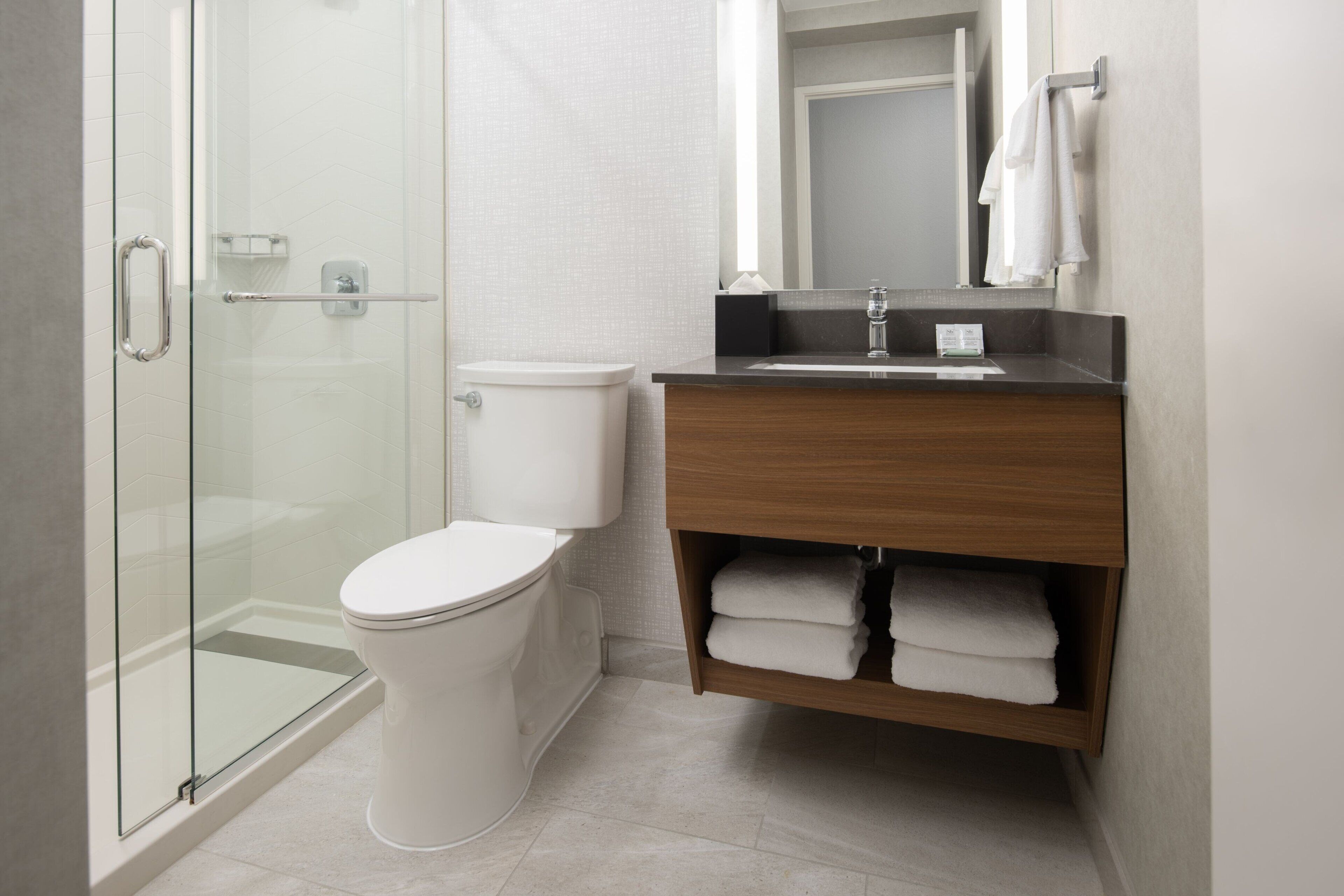 A modern bathroom with a glass shower enclosure, a white toilet, and a wooden vanity with a mirror and towel rack.