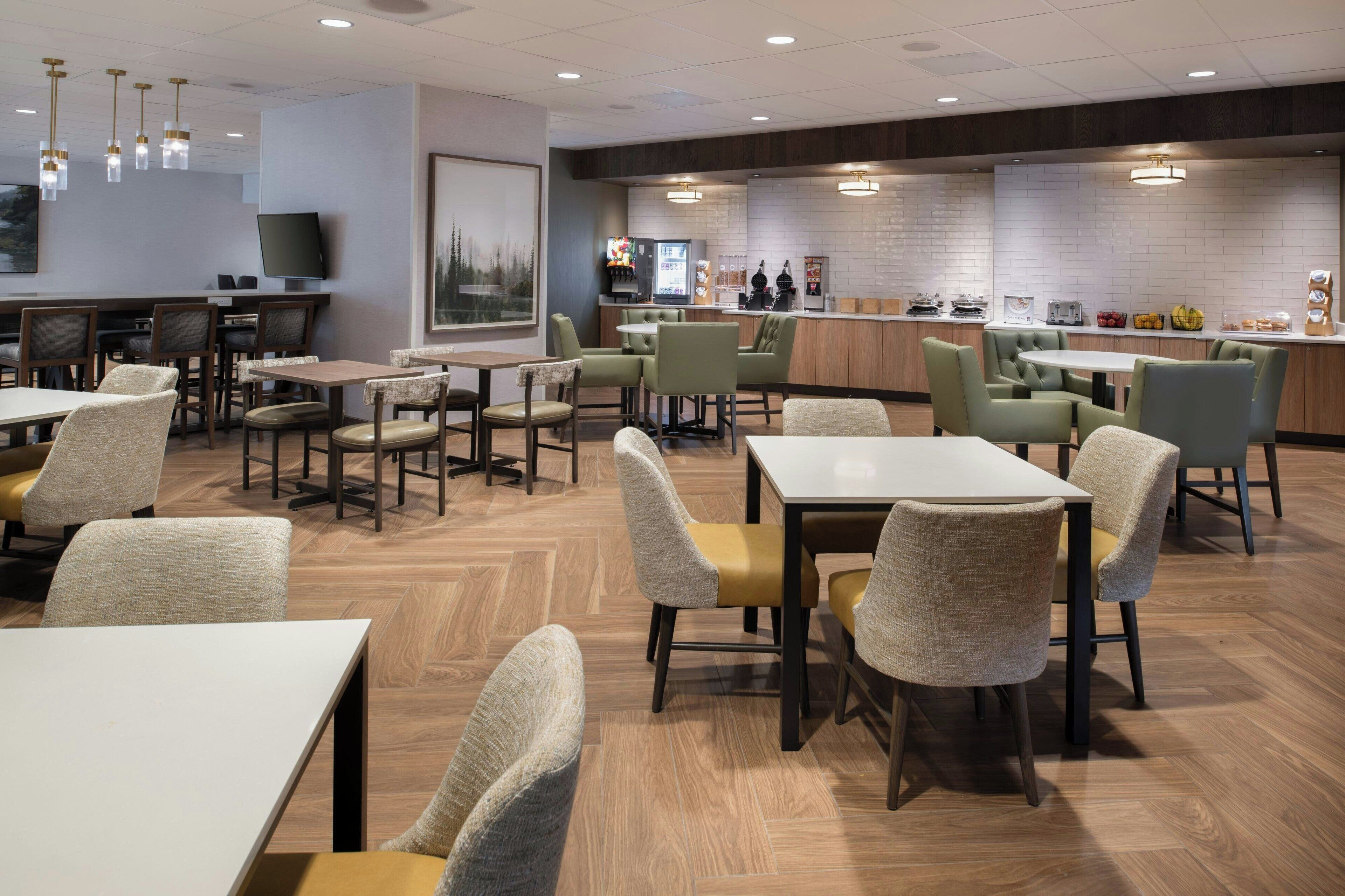 A modern restaurant with multiple dining tables and chairs, a bar area, and a kitchen in the background.