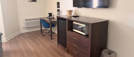 Una habitación compacta con escritorio, silla, kitchenette y televisor de pared.