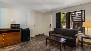 A living room with a dark brown sofa, a wooden coffee table, a small black refrigerator, a wooden cabinet with drawers, and a window with curtains.