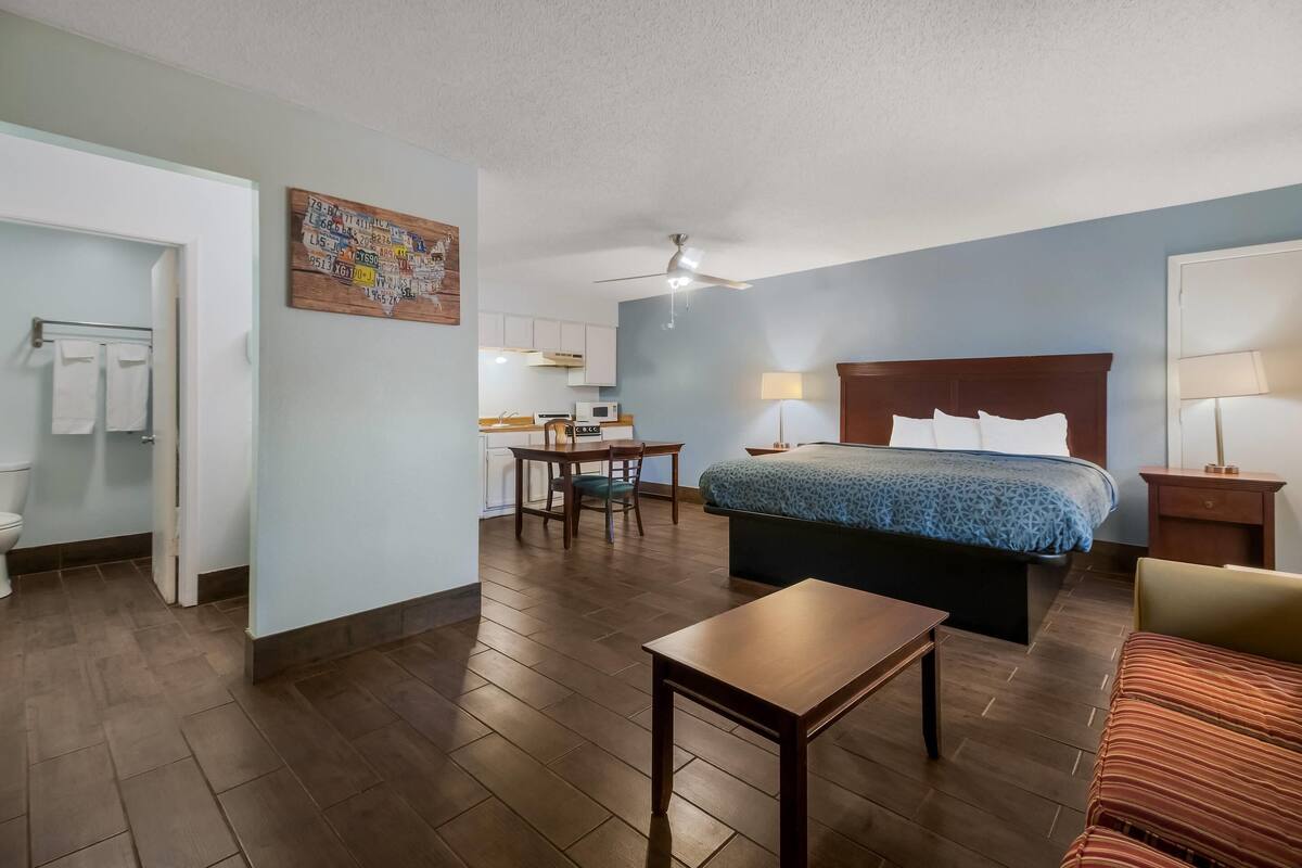 A hotel room with a bed, a desk, a chair, a small table, a nightstand, a lamp, and a bathroom with a toilet and towel rack.
