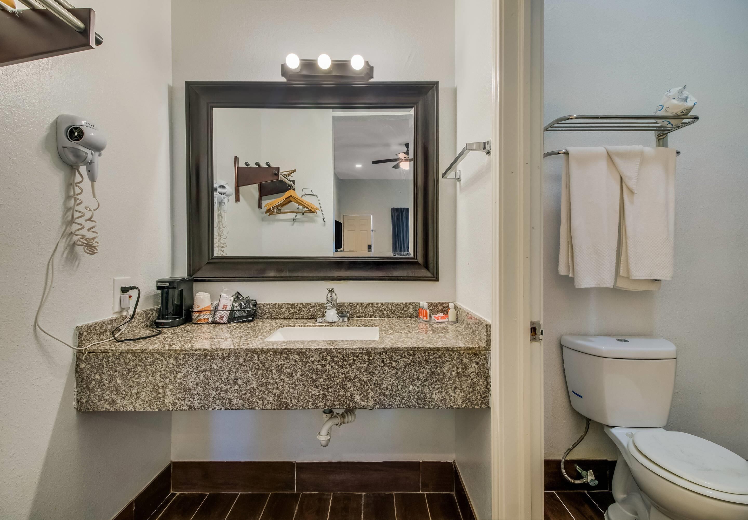A bathroom with a granite countertop sink, a mirror with lights, a hairdryer, a towel rack, and a toilet.