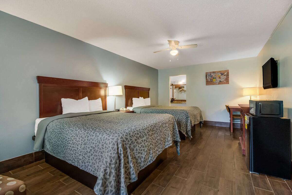 A hotel room with two beds, a ceiling fan, a desk with a chair, a television, and a refrigerator.