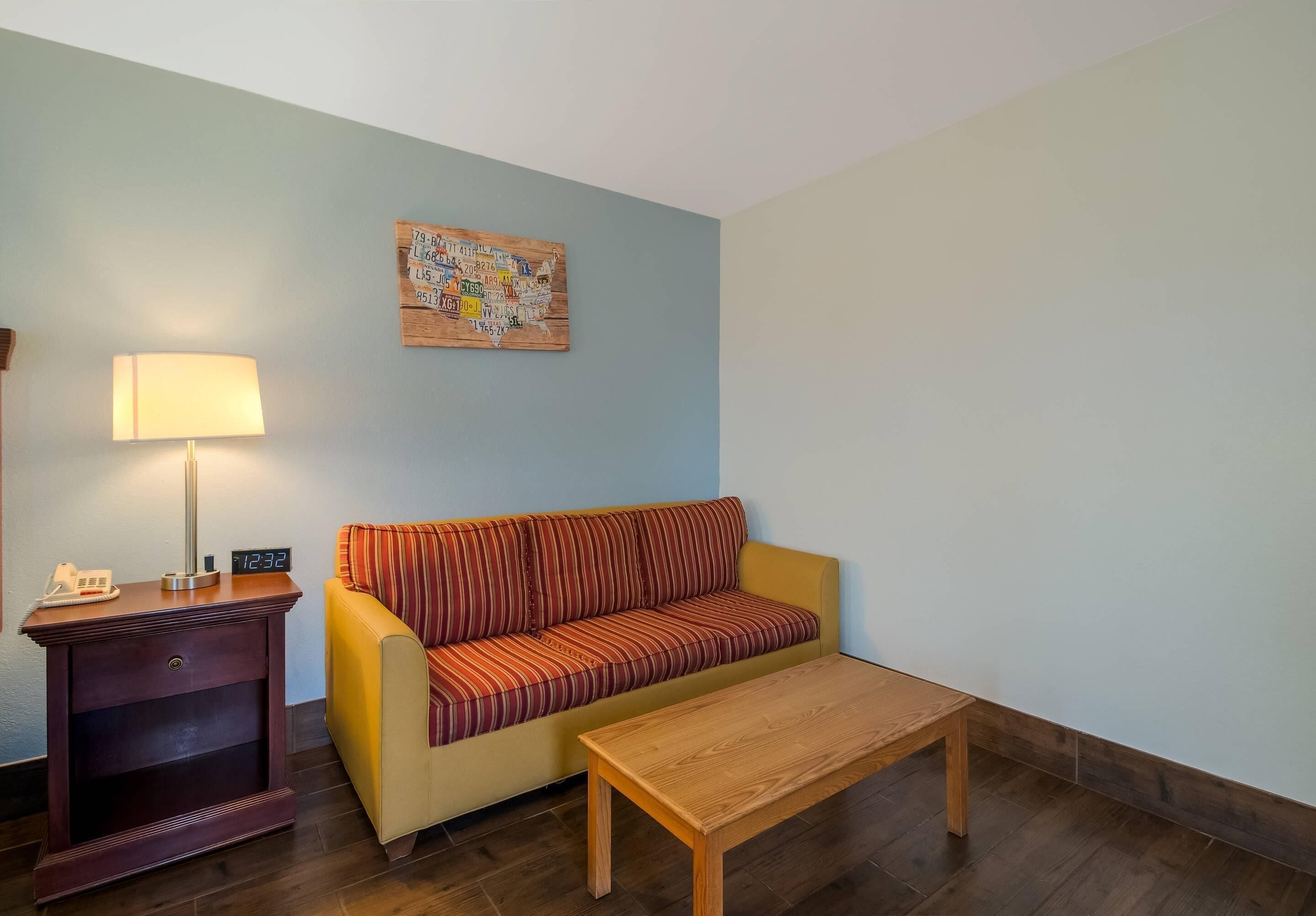 A living room with a yellow sofa, a wooden coffee table, a lamp, a clock, and a wall art piece.