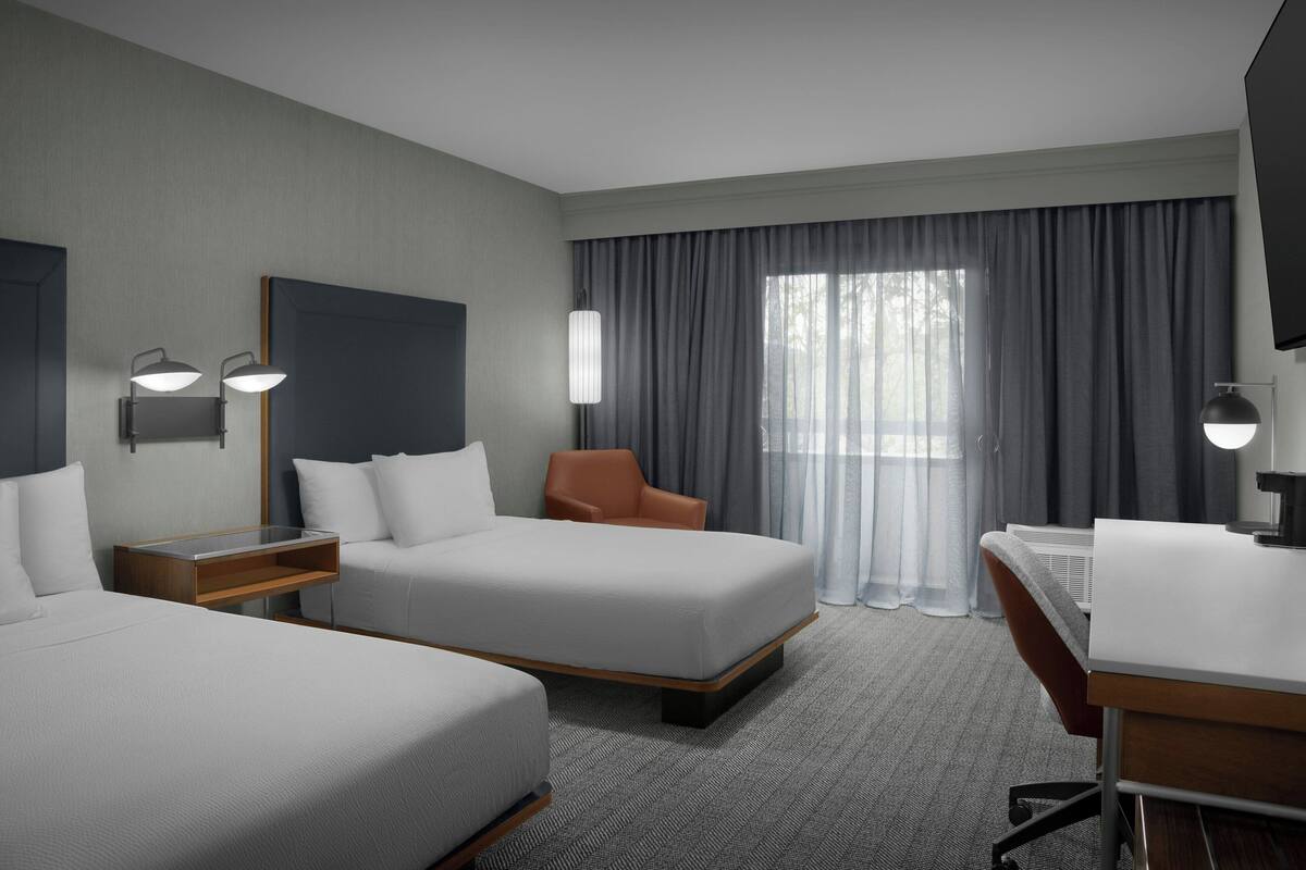Habitación de hotel con dos camas, un escritorio, una silla, un televisor y un ventanal grande con cortinas.