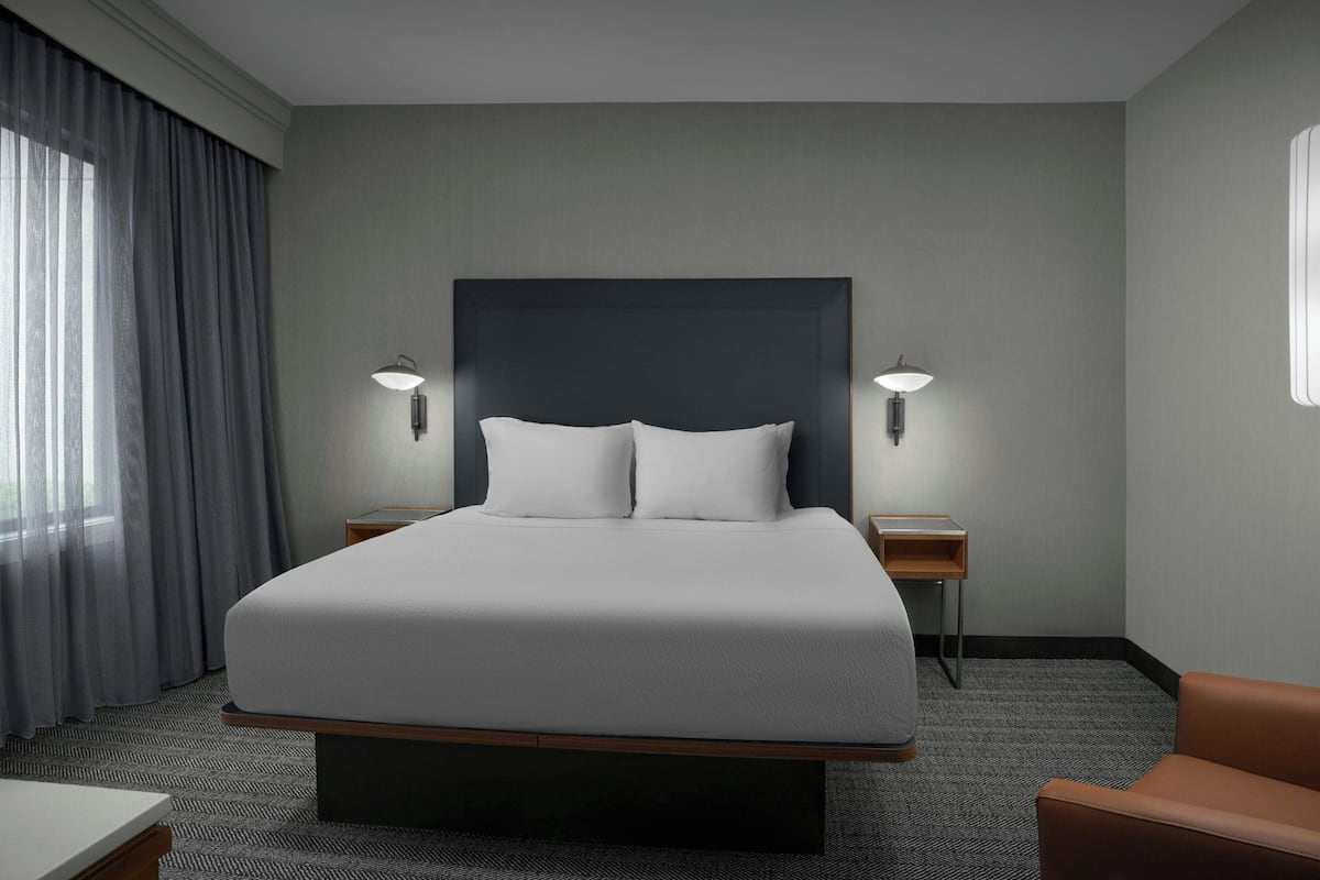 Una habitación de hotel con una cama grande, dos lámparas de noche, una mesita pequeña, una silla y una ventana con cortinas.