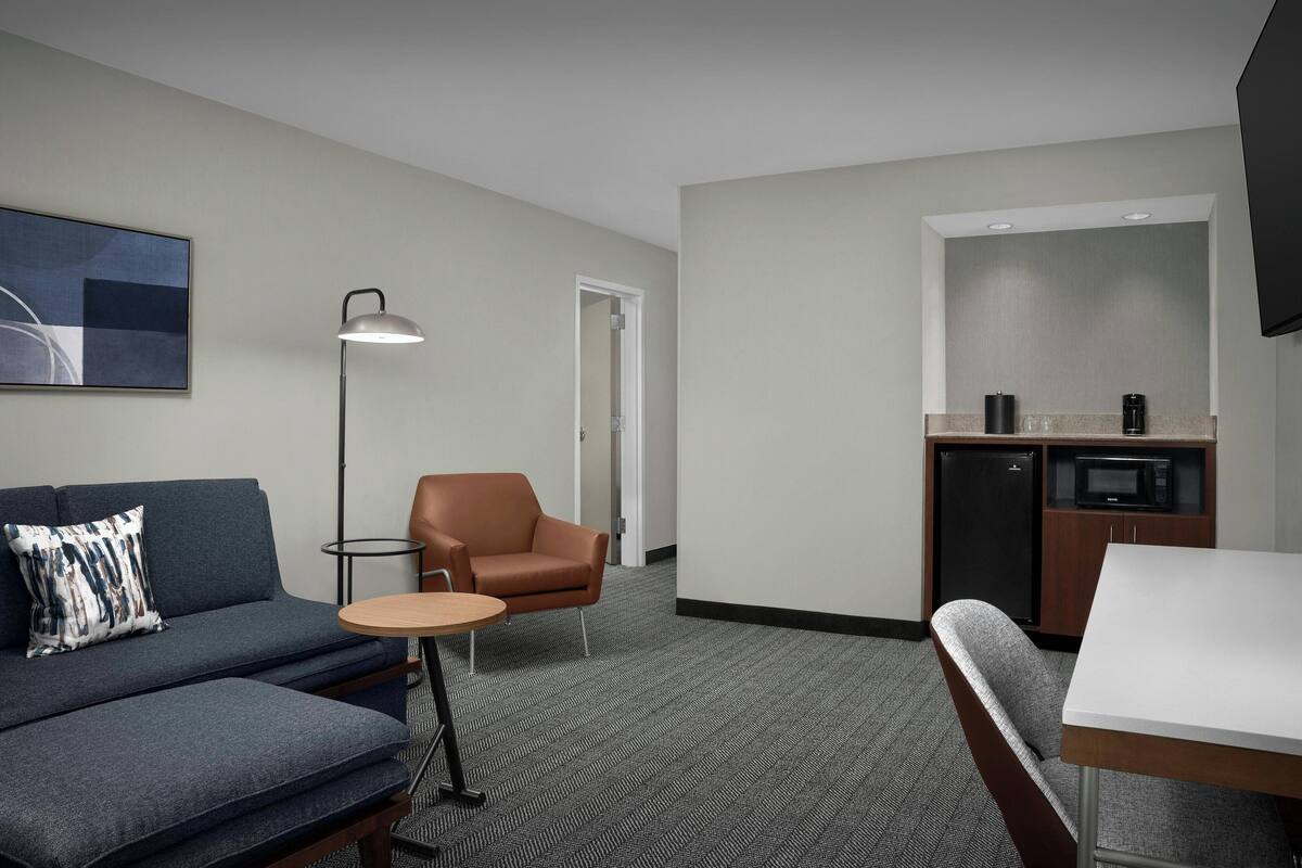 A hotel room with a sofa, armchair, small round table, and a kitchenette area.