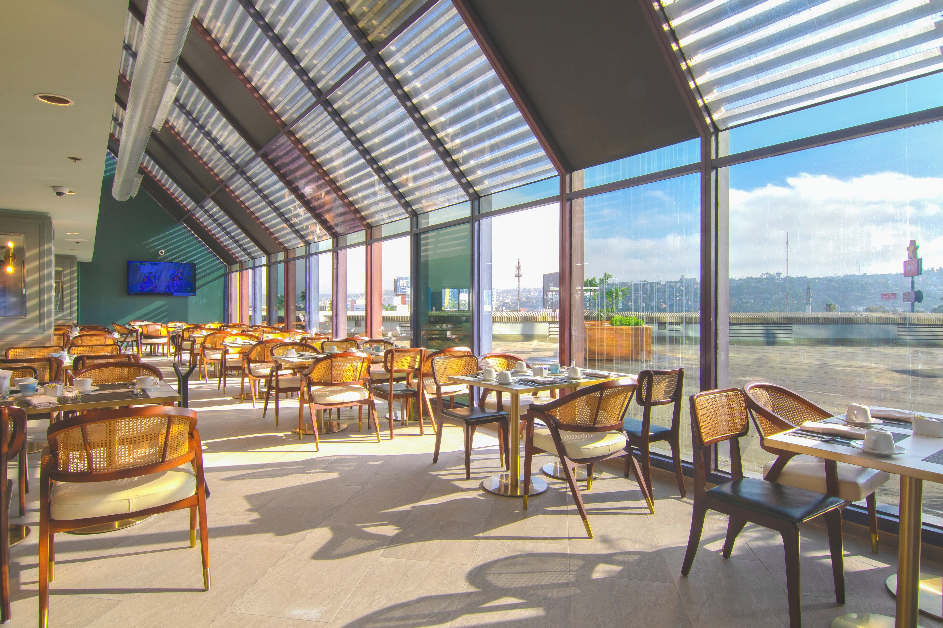 A spacious indoor dining area with multiple tables and chairs, large windows offering a view of the cityscape, and a flat-screen TV mounted on the wall.