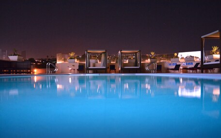 A nighttime swimming pool with lounge chairs and umbrellas, surrounded by buildings and city lights.