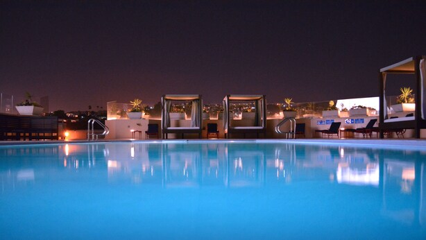 Piscina à noite com espreguiçadeiras e guarda-sóis, rodeada por edifícios e luzes da cidade.